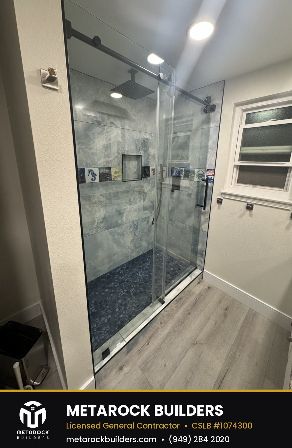 Shower remodel in Orange County by Sunward Builders Inc., completed after photo with blue-gray onyx-look tile, pebble mosaic floor, matte black hardware, and sliding glass door